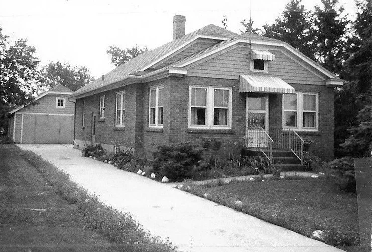 Black-and-white photo of a single-story brick bungalow with a front porch and awning, concrete driveway leading to a detached garage, and trees in the yard.