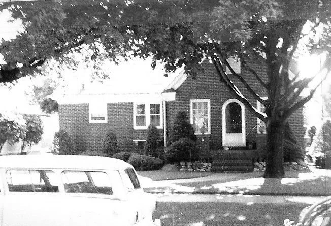 Black-and-white photo of a single-story brick house with an arched front door, large shade tree in the yard, and a vintage station wagon parked on the street.