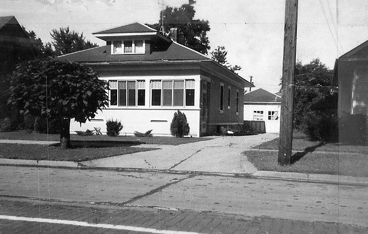 Black-and-white photo of a one-story bungalow with an enclosed front porch, driveway leading to a detached garage, a small tree and a utility pole in the foreground.