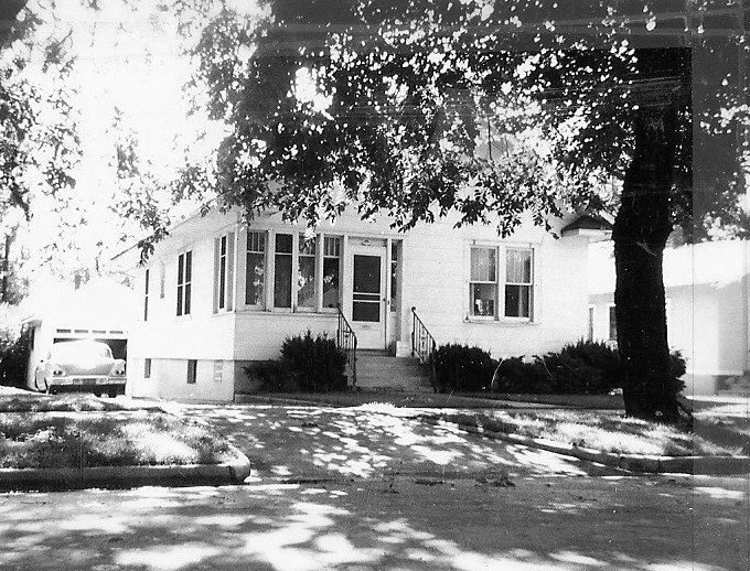 Black-and-white photo of a one-story house with a small front porch and steps, shrubs along the foundation, a large tree casting dappled shadows, and a car parked in the driveway.