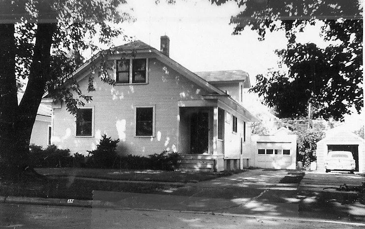 Black-and-white photo of a two-story clapboard house with a small front porch, driveway leading to two detached garages, and a car parked in the right garage, shaded by large trees.