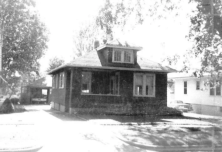 Black-and-white photo of a one-story brick bungalow with a central dormer, covered front porch, and trees along a sunny street.