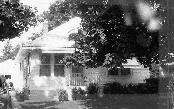 Black-and-white photo of a small single-story house with a shaded front porch, large tree, and the number "261" by the steps.