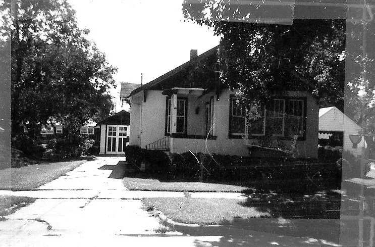 Black-and-white photo of a small single-story bungalow with front steps, shrubs, driveway and large trees casting shadows.