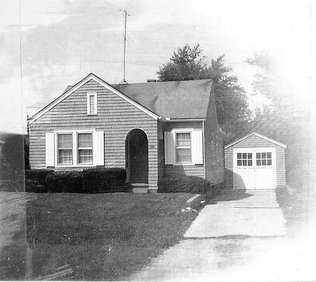 Black-and-white vintage photo of a small single-story house with shingle siding, arched front entry, lawn, driveway and detached garage.