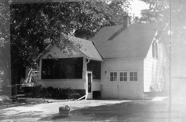 Black-and-white photo of a small house with a covered front porch, attached two-car garage, trees overhead and a planter in the driveway.