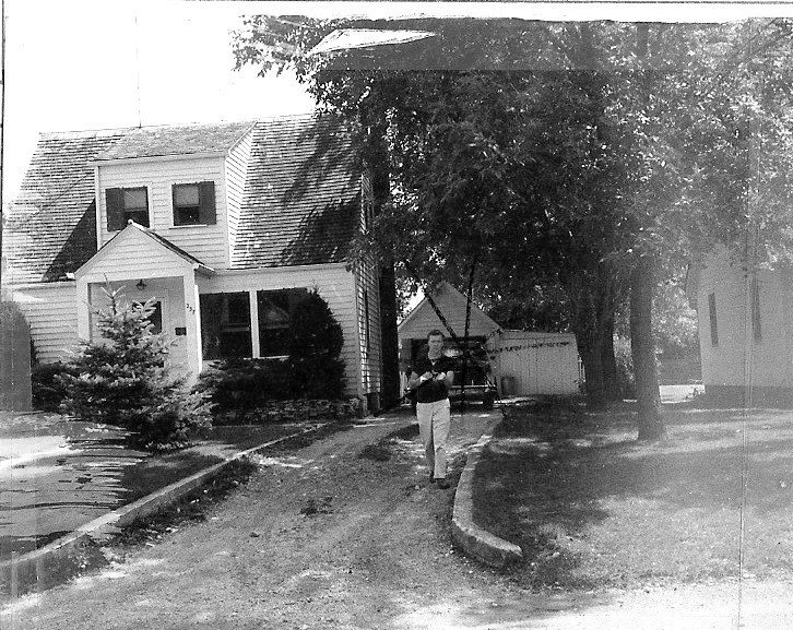 Black-and-white photo of a two-story house with trees and a person walking down the driveway toward the camera.