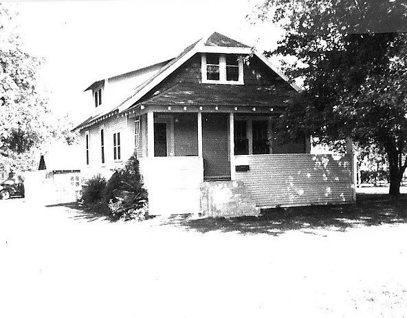 Black-and-white photo of a small bungalow with a covered front porch, dormer window, and trees at the sides.