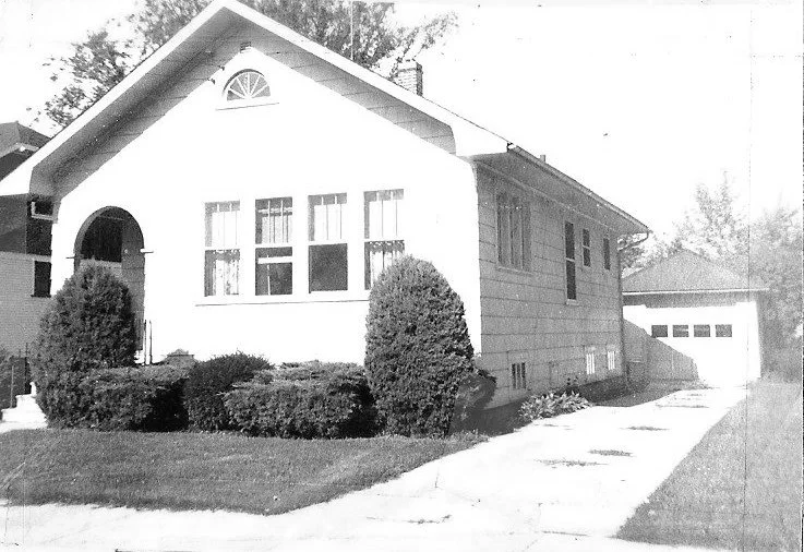 Black-and-white photo of a single-story bungalow with an arched front porch, large front windows, trimmed shrubs, and a driveway leading to a detached garage.