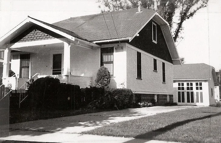 Black-and-white photo of a single-story bungalow with a covered front porch, shrubs along the foundation, a driveway leading to a detached garage, and a steep side roof.
