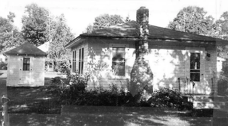 Black-and-white photo of a small single-story house with a central brick chimney, a front porch, and a small outbuilding to the left, set among trees and lawn.
