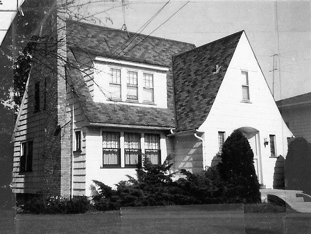 Black-and-white photo of a two-story suburban house with steep gabled roof, chimney, arched front entry, and shrubs in the yard.