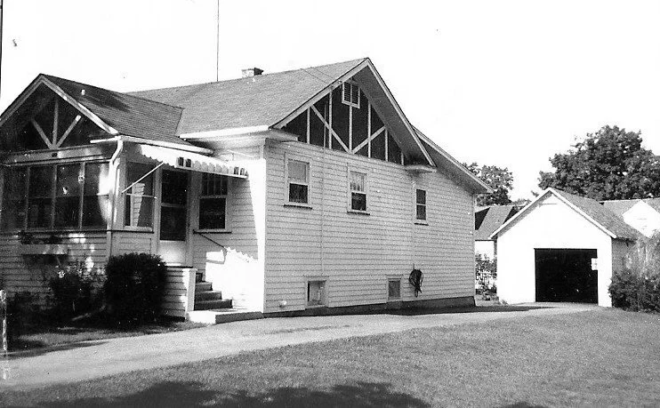 Black-and-white photo of a single-story bungalow with a small enclosed porch, driveway, lawn, and a detached garage.