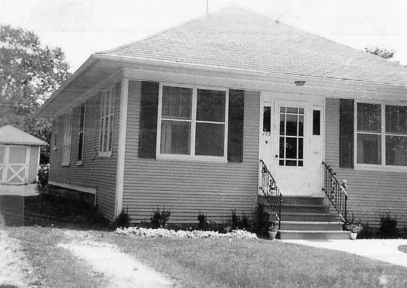 Black-and-white photo of a single-story bungalow with front steps and storm door, two large front windows, small shed at left, and a grassy yard.