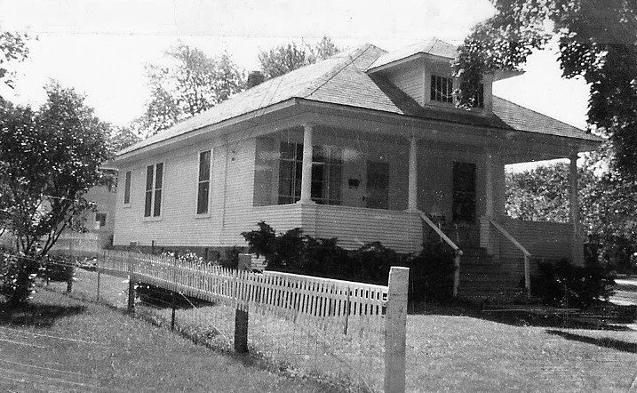 Black-and-white photo of a single-story wood-frame bungalow with a covered front porch, steps, white picket fence and trees around the yard.