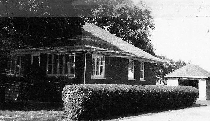 Black-and-white photo of a one-story brick house with a low trimmed hedge in front and a detached garage to the right.