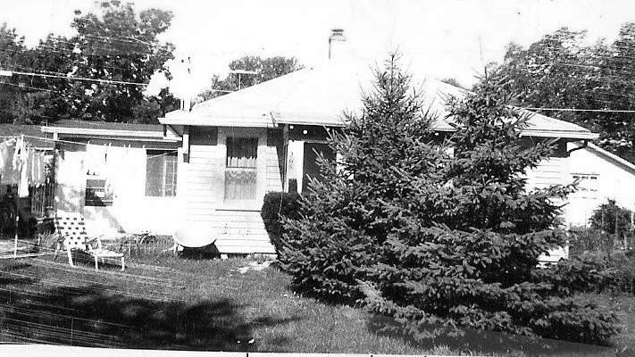 Black-and-white photo of a small single-story house with two large evergreen trees in the front yard, a lawn with a folding chair, and a clothesline with laundry.