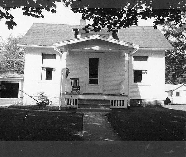 Black-and-white photo of a small single-story house with a covered front porch, a wooden chair on the steps, and a walkway leading to the door.