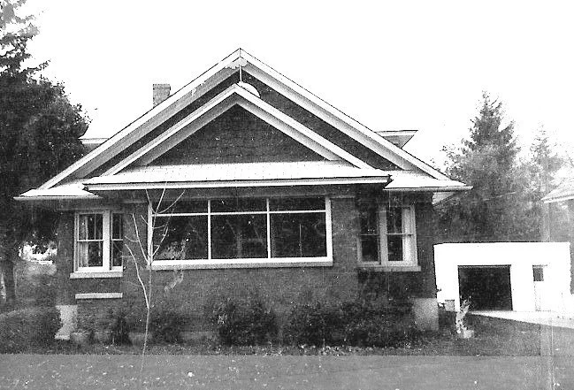 Black-and-white photo of a one-story brick bungalow with a large front window, gabled roof, small yard, and a detached garage to the right.