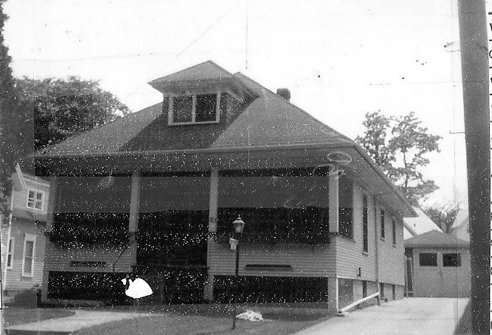 Black-and-white photo of a bungalow-style house with a wide front porch supported by columns, a dormer window, driveway leading to a detached garage, and a lamppost in the yard.