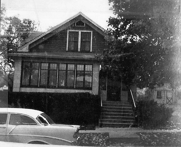 Black-and-white photo of a two-story bungalow with an enclosed front porch, concrete steps leading to the entrance, trees at the side, and a vintage 1950s car parked on the street in front.