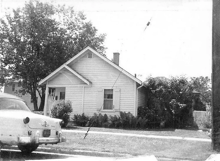 Black-and-white photo of a small single-story clapboard house with a front lawn and tree, and a vintage car parked in the foreground.
