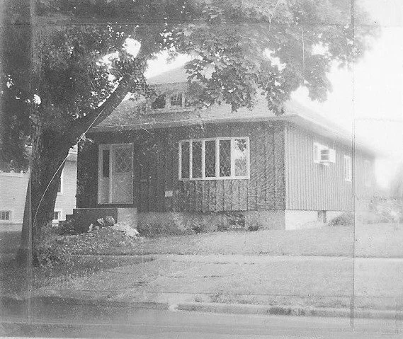 Black-and-white photo of a single-story wood-paneled house with a front porch, large bay window, and a tree in the front yard.