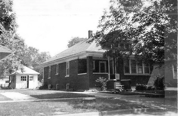 Black-and-white photo of a single-story brick bungalow with a small front porch and steps, a large tree shading the yard, and a detached garage to the left.