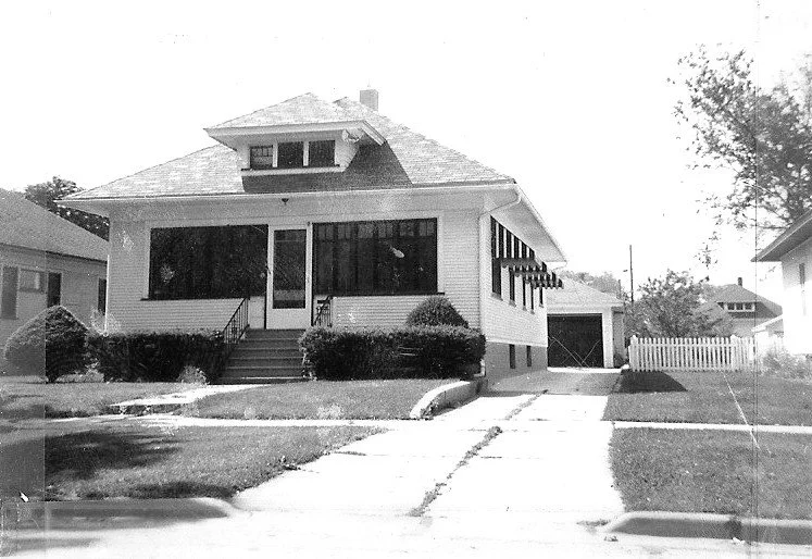 Black-and-white photo of a single-story bungalow with an enclosed front porch, steps and shrubs, a driveway leading to a detached garage, and a small lawn with a white picket fence.