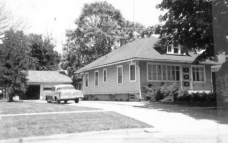 Black-and-white photo of a mid-20th-century bungalow with a detached garage and a vintage car parked in the driveway beneath leafy trees.