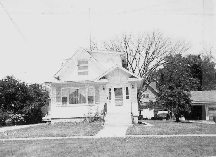 Black-and-white photo of a two-story bungalow with front steps and porch, a bare tree behind it, lawn in front, and a garage and car to the right.