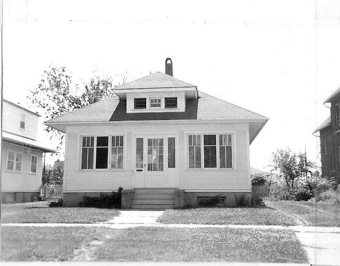 Front-facing black-and-white photo of a single-story bungalow with an enclosed front porch, central door and steps, a small dormer and chimney, and neighboring houses on each side.
