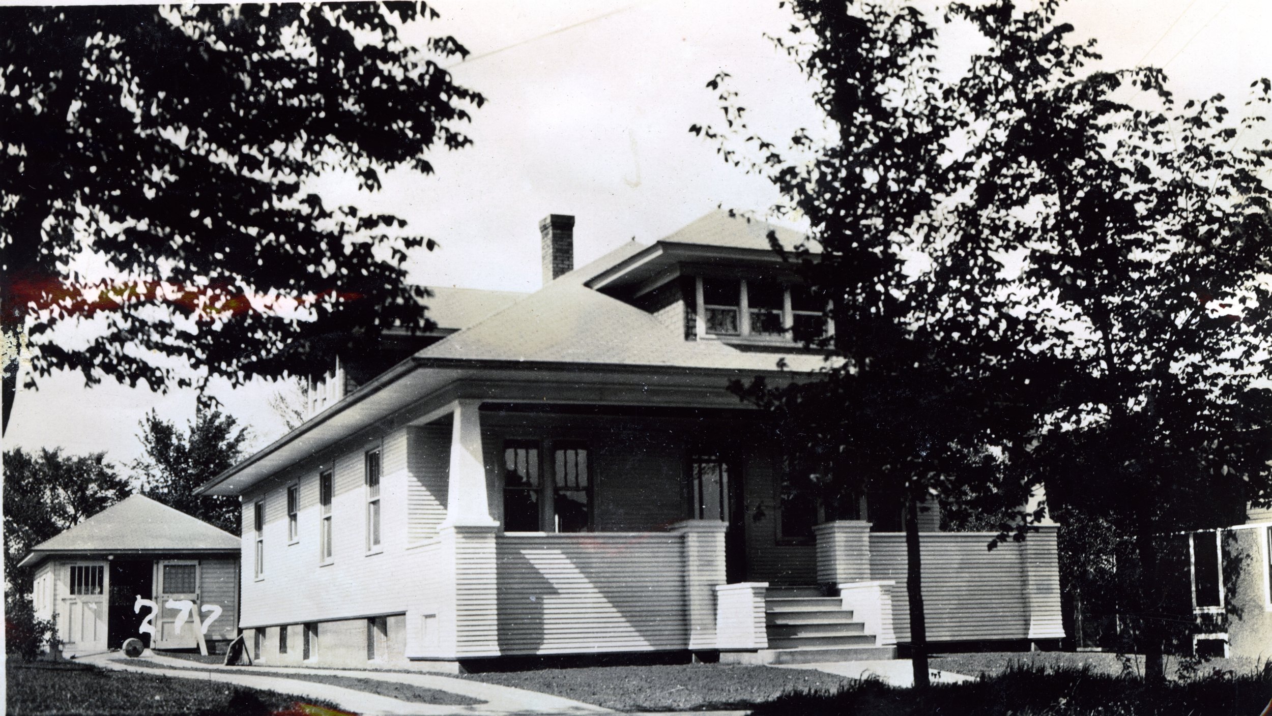 Black-and-white photo of a two-story Craftsman house with a covered front porch, detached garage at left labeled "272", and large trees partially obscuring the roof.