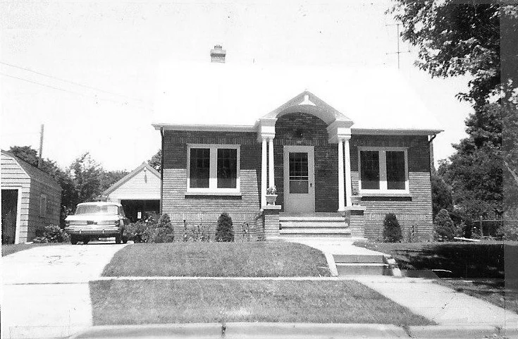 Black-and-white photo of a small brick bungalow with a central front door and steps, two windows flanking the entrance, a driveway leading to a detached garage, and a vintage car parked to the left.