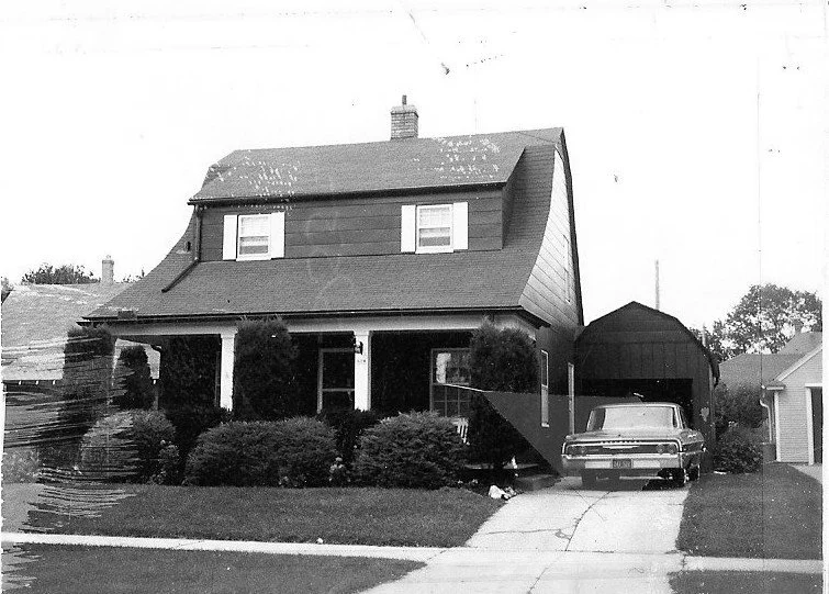 Black-and-white photo of a two-story house with a covered front porch, shrubs lining the yard, and a vintage car parked in the driveway beside a detached garage.