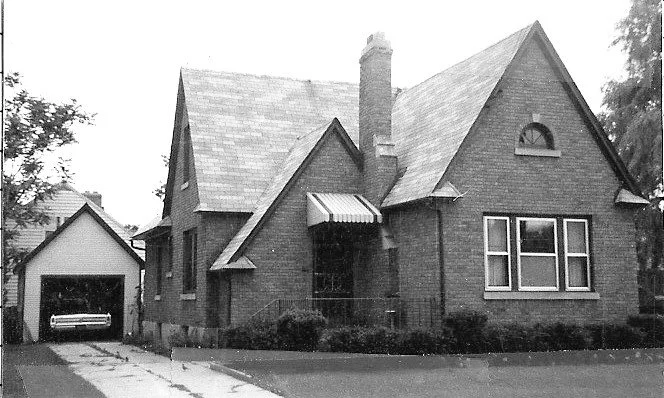 Black-and-white photo of a one-story brick cottage with steep gabled roof, central chimney, awning over the front entry, shrubs along the foundation, and a detached garage with a car.