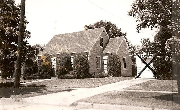 Black-and-white photo of a small gable-roof house with tall shrubs along the front and a driveway leading to a detached garage marked with a large X.