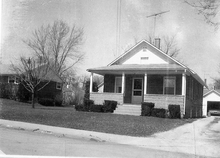 Black-and-white photo of a single-story bungalow with a covered front porch, low shrubs, driveway leading to a detached garage, and leafless trees in the yard.