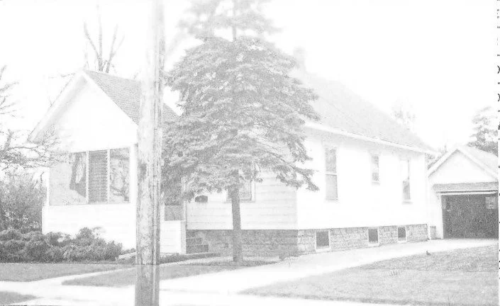 Small single-story white house with a covered front porch, a large evergreen tree in front, a sidewalk, utility pole, and a detached garage.