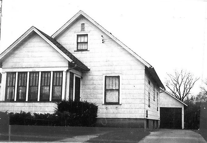 Black-and-white photo of a single-story gabled house with an enclosed front porch, shrubs, and a driveway leading to a detached garage.