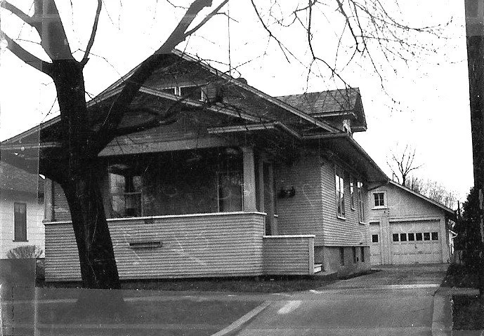 Black-and-white photo of a single-story bungalow with a covered front porch and detached two-car garage, bare trees in front, and a street curb in the foreground.