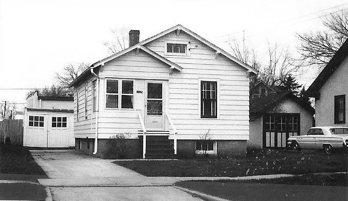 Black-and-white photo of a small single-story white bungalow with front steps, a detached garage at left, and a vintage car parked by the neighboring driveway.