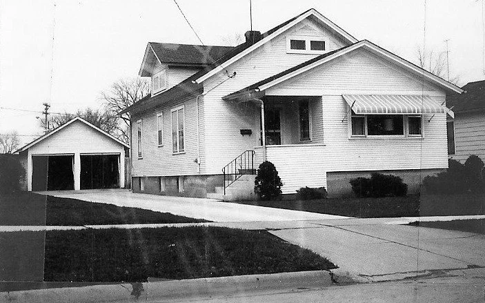 Black-and-white photo of a single-story bungalow with a front porch, concrete driveway, and a detached two-car garage.