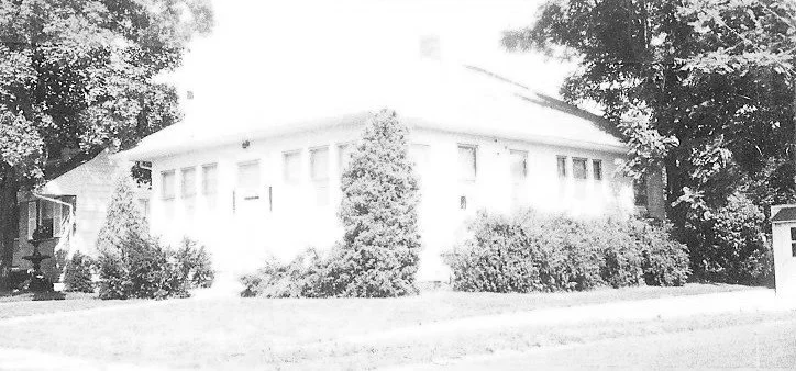 Black-and-white overexposed photo of a single-story house with a row of tall shrubs and trees along the front.