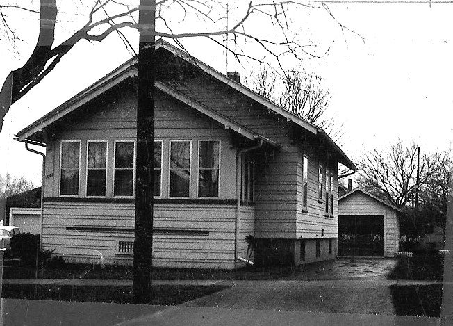 Black-and-white photo of a one-story bungalow with an enclosed front porch, driveway leading to a detached garage, and bare trees in the yard.
