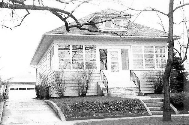 Black-and-white photo of a single-story bungalow with an enclosed front porch, concrete steps and metal railings, driveway leading to a detached garage, and leafless trees in the yard.