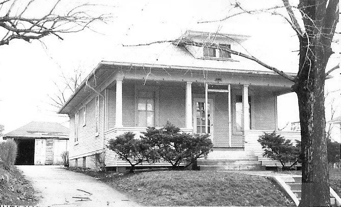 Black-and-white photo of a single-story bungalow with a front porch, shrubs, driveway, and detached garage.
