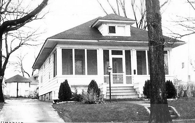 Black-and-white photo of a single-story bungalow with a screened front porch, central steps and lamppost, driveway to a detached garage.