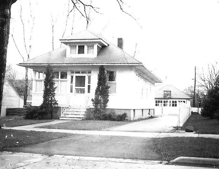 Black-and-white photo of a single-story bungalow with a front enclosed porch, steps flanked by two tall shrubs, and a driveway leading to a detached garage.