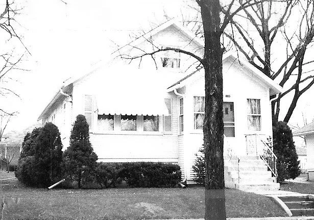 Black-and-white photo of a small bungalow-style house with front steps and awning, flanked by leafless trees and trimmed shrubs on a residential lawn.